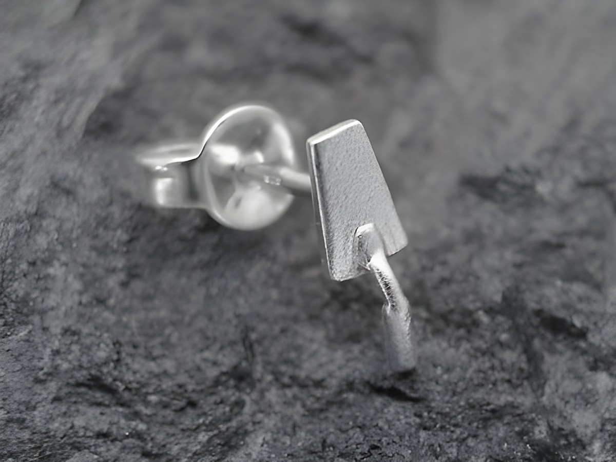 Ein einzelner Ohrring in Form einer Maurerkelle aus Sterling Silber, hergestellt von Juwelier Harnisch, auf einem neutralen Untergrund abgebildet.