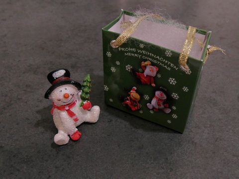 Ein Schneemann sitzt neben einer Geschenkbox mit weihnachtlichen Motiven. Die Box ist grün und trägt die Aufschrift Frohe Weihnachten. Hersteller ist Juwelier Harnisch.