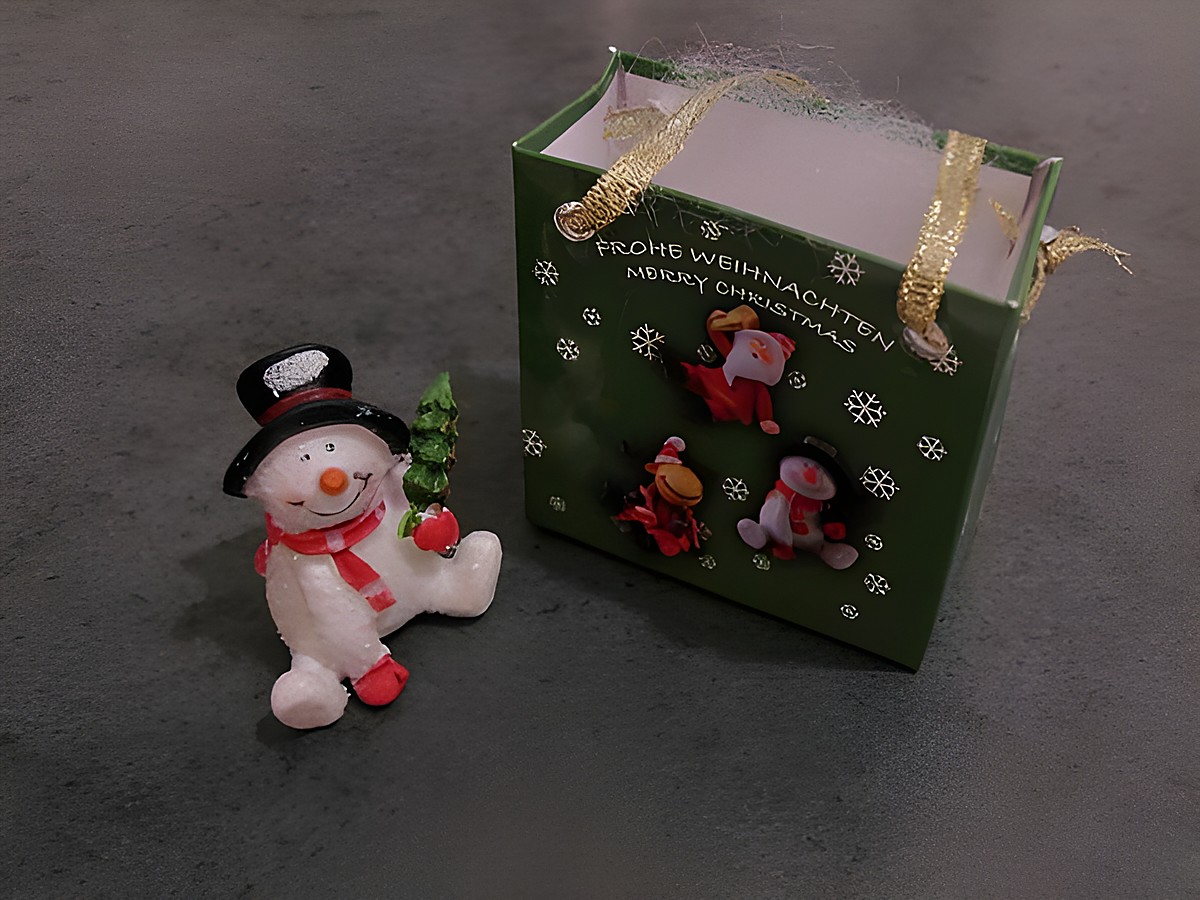 Ein Schneemann sitzt neben einer Geschenkbox mit weihnachtlichen Motiven. Die Box ist grün und trägt die Aufschrift Frohe Weihnachten. Hersteller ist Juwelier Harnisch.