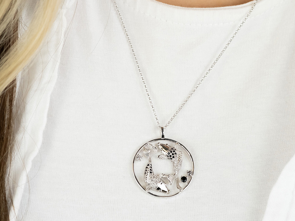 Silberne Kette mit Yin Yang Fische Anhänger, verziert mit Zirkonia. Der Anhänger zeigt zwei stilisierte Fische in einer runden Form. Trage hierzu ein schlichtes weißes Oberteil.