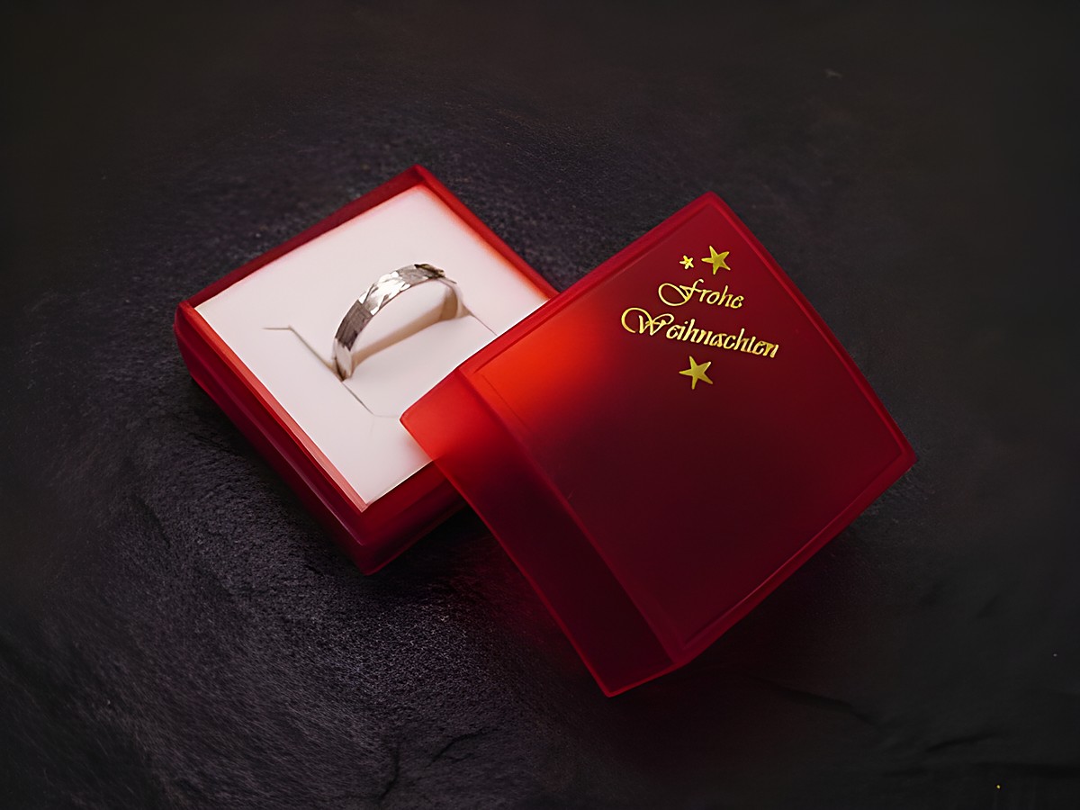 Eine rote Geschenkschachtel mit der Aufschrift Frohe Weihnachten öffnet sich und zeigt einen silbernen Ring im Innenraum, Hersteller Juwelier Harnisch, Größe 4,5 cm, ideal für Weihnachten.