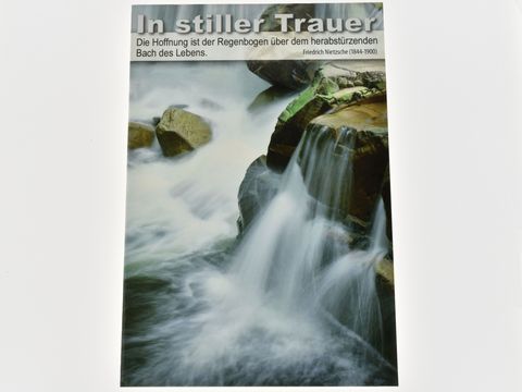 Trauerkarte mit einem sanften Wasserfall, umgeben von Steinen. Oben der Schriftzug In stiller Trauer und ein Zitat von Friedrich Nietzsche. Hersteller Juwelier Harnisch.