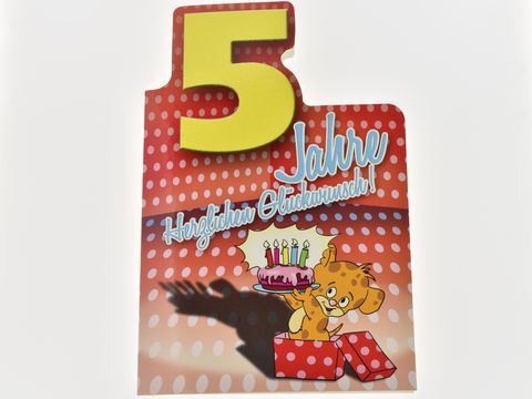 Geburtstagskarte mit einem fröhlichen Bärchen, das eine Torte mit Kerzen auf einem roten Hintergrund mit weißen Punkten präsentiert. Die Karte ist für einen fünften Geburtstag gestaltet. Hersteller: Juwelier Harnisch.
