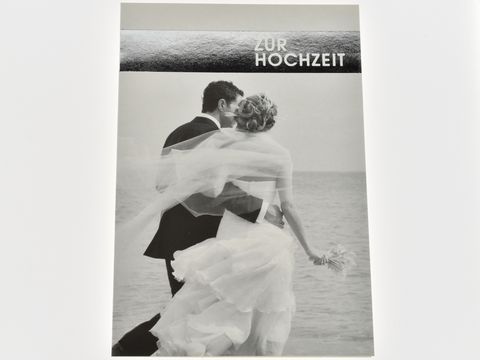 Hochzeitskarte mit einem Brautpaar am Meer in Schwarz-Weiß, der Bräutigam hält die Braut, die ein fließendes Kleid trägt, im Hintergrund ist das Wasser zu sehen, Hersteller Juwelier Harnisch.