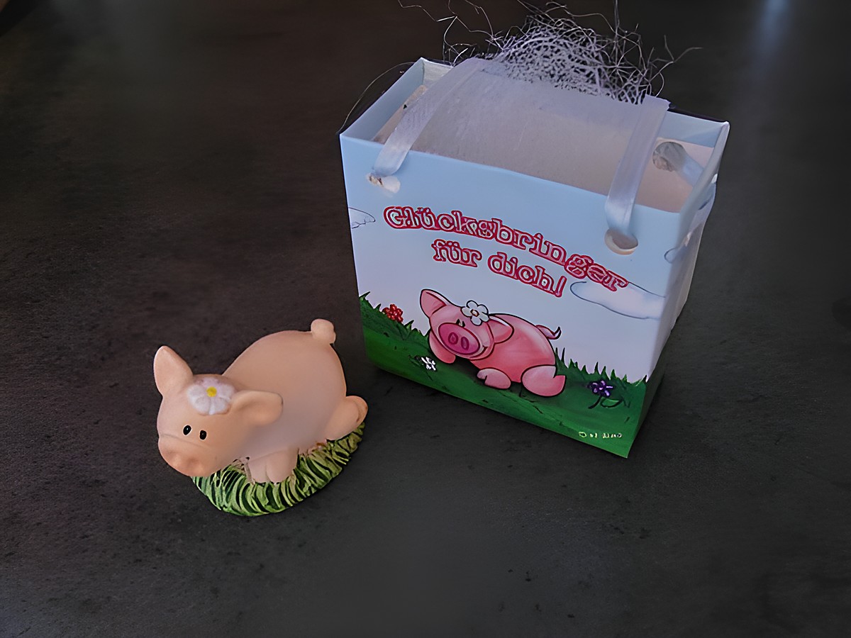 Glücksschweinchen mit Geschenktasche liegend, Hersteller Juwelier Harnisch, dekorative Figur mit blühendem Gras und einer Geschenktasche in fröhlichen Farben.