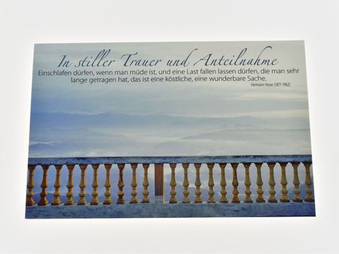 Eine Trauerkarte mit dem Titel "In stiller Trauer und Anteilnahme", die in sanften Blau- und Grautönen gehalten ist. Im Hintergrund ist eine Landschaft mit einem Geländer zu sehen und eine ruhige, melancholische Atmosphäre vermittelt.