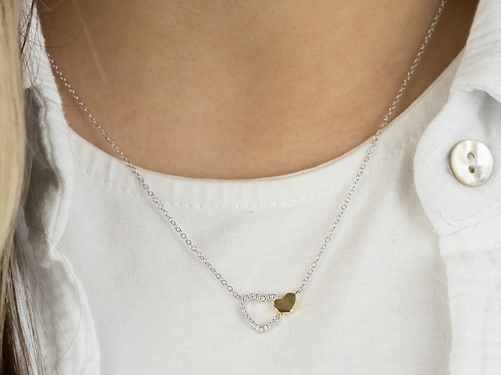 Wunderschöne Engelsrufer ERN-HEARTBEAT-ZI-BIG Kette in Bicolor mit zwei Herzen, eines mit Zirkonia verziert, an einer zarten Silberkette, getragen über einem weißen Oberteil.