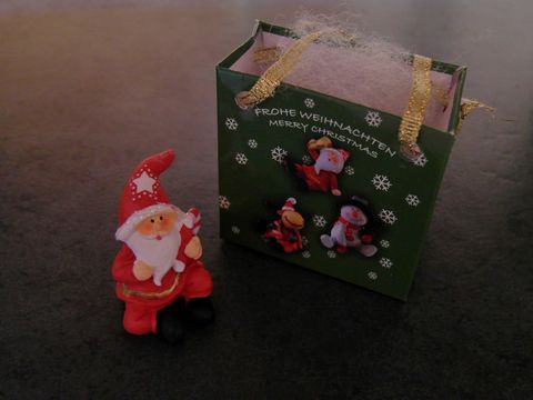 Weihnachtsmann mit Geschenktasche sitzend, hält eine Zuckerstange in der Hand, neben einer grünen Geschenktüte mit festlichem Design von Juwelier Harnisch, für Weihnachten.
