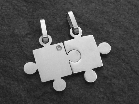 Zwei Partneranhänger in Form von Puzzlestücken aus Edelstahl mit einem Kristall. Die Anhänger sind poliert und zeigen eine moderne, schlichte Gestaltung, hergestellt von Juwelier Harnisch.
