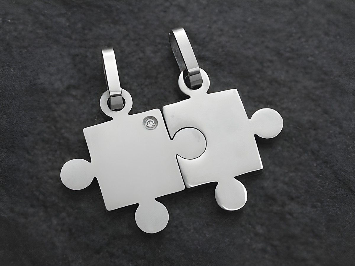 Zwei Partneranhänger in Form von Puzzlestücken aus Edelstahl mit einem Kristall. Die Anhänger sind poliert und zeigen eine moderne, schlichte Gestaltung, hergestellt von Juwelier Harnisch.
