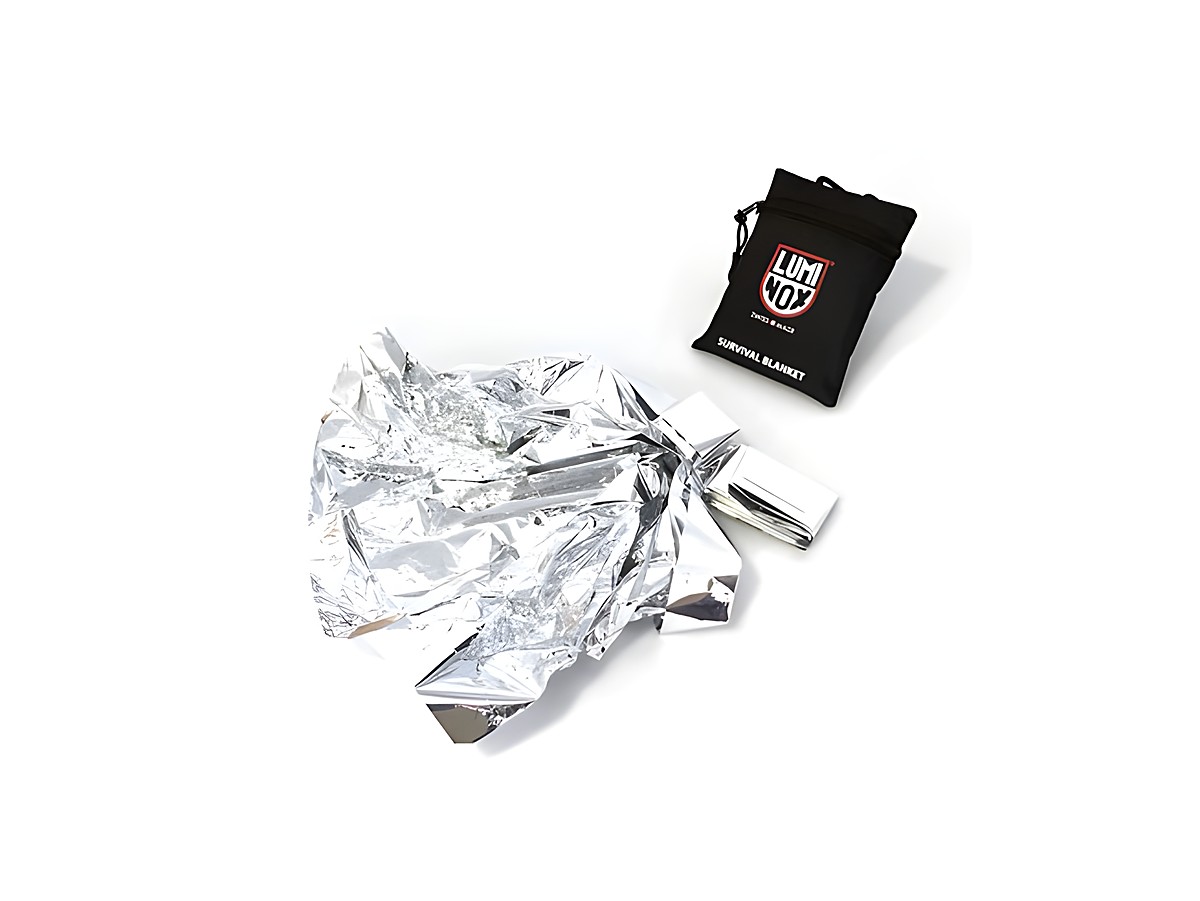Die Luminox Survival Decke in silberner Farbe liegt zusammengefaltet auf weißem Hintergrund, neben ihr befindet sich die schwarze Verpackung mit Luminox Logo und der Bezeichnung Survival Blanket.