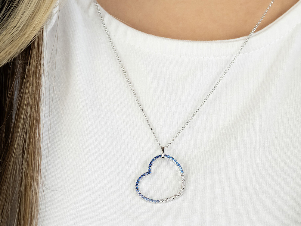 Silberne Kette mit einem herzförmigen Anhänger, verziert mit blauen und klaren Zirkonia, getragen von einer Person mit heller Kleidung und langen Haaren.
