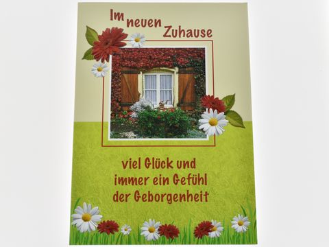Eine liebevoll gestaltete Grußkarte mit dem Titel "Im neuen Zuhause". Im Hintergrund ist ein Fenster mit Efeu und bunten Blumen. Unter dem Fenster steht der Text "viel Glück und immer ein Gefühl der Geborgenheit". 