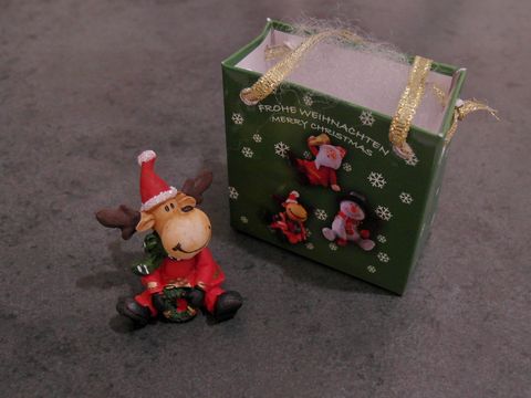 Elch im Weihnachtsmannkostüm sitzt mit einem Tannenkranz und einer Geschenktasche neben sich. Hersteller ist Juwelier Harnisch, ideal für festliche Dekoration.