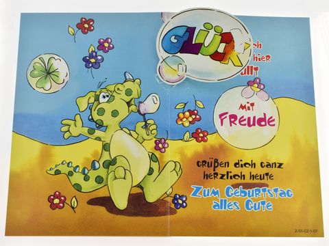 Lustige Geburtstagskarte mit einem niedlichen, grünen Dino, der fröhlich Seifenblasen bläst und von bunten Blumen umgeben ist. Bunte Schrift mit herzlichen Geburtstagsgrüßen.