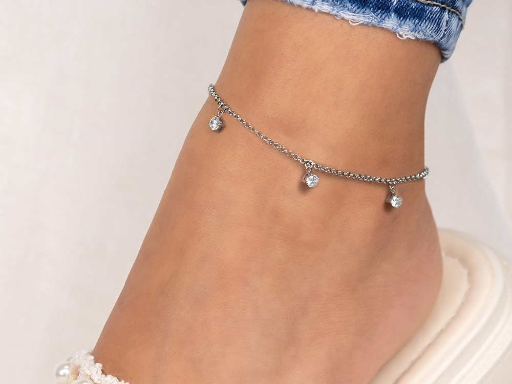 Fußkette aus Edelstahl mit Zirkonia in glänzender Ausführung, ergänzt durch kleine, funkelnde Anhänger. Die Kette hat eine Gesamtlänge von 22 plus 5 cm und wird am Knöchel getragen.