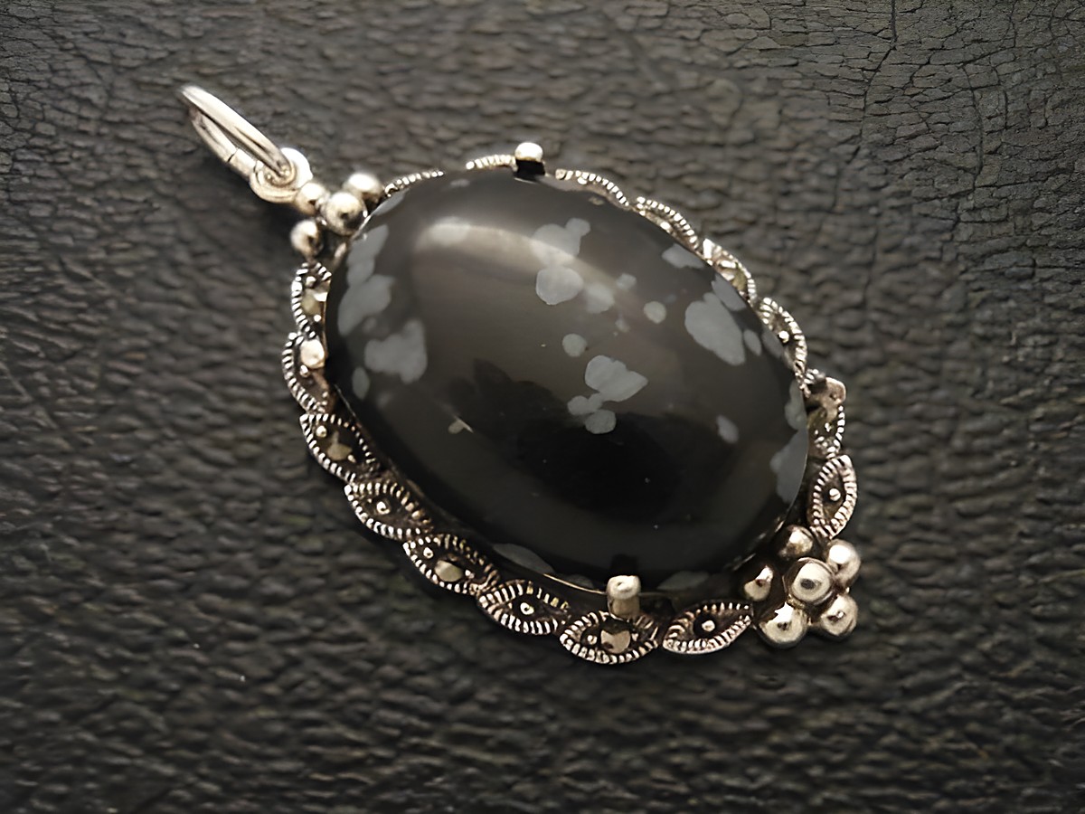 Schneeflocken-Obsidian Cabochon in einem filigranen Silbermedaillon Anhänger, elegant gestaltet mit geschwungenen Verzierungen und kleinen Details, ideal als Schmuckstück für besondere Anlässe.