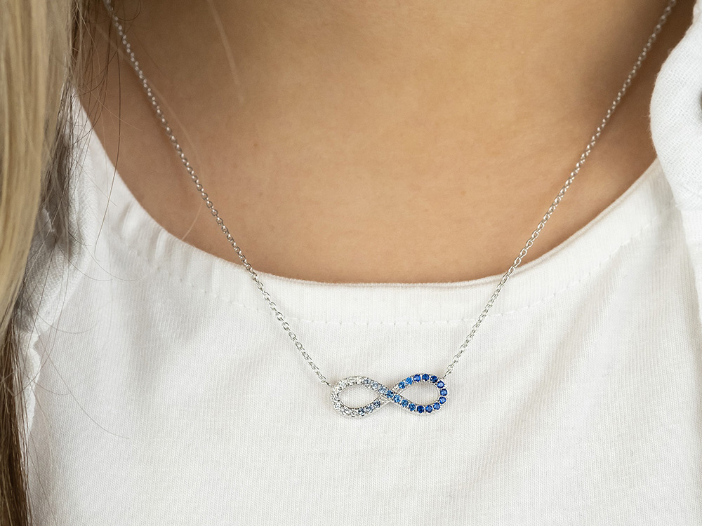 Silberne Kette mit Unendlichkeitssymbol, besetzt mit Zirkonia, getragen von einer Person. Das Schmuckstück hat eine Länge von 40 plus 4 cm und zeigt blaue und klare Steine.