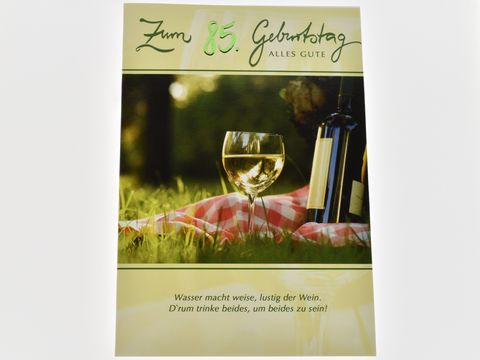 Eine Geburtstagskarte mit der Aufschrift zum 85. Geburtstag alles Gute, einer Flasche Wein und einem gefüllten Weinglas auf einer Picknickdecke im Grünen.