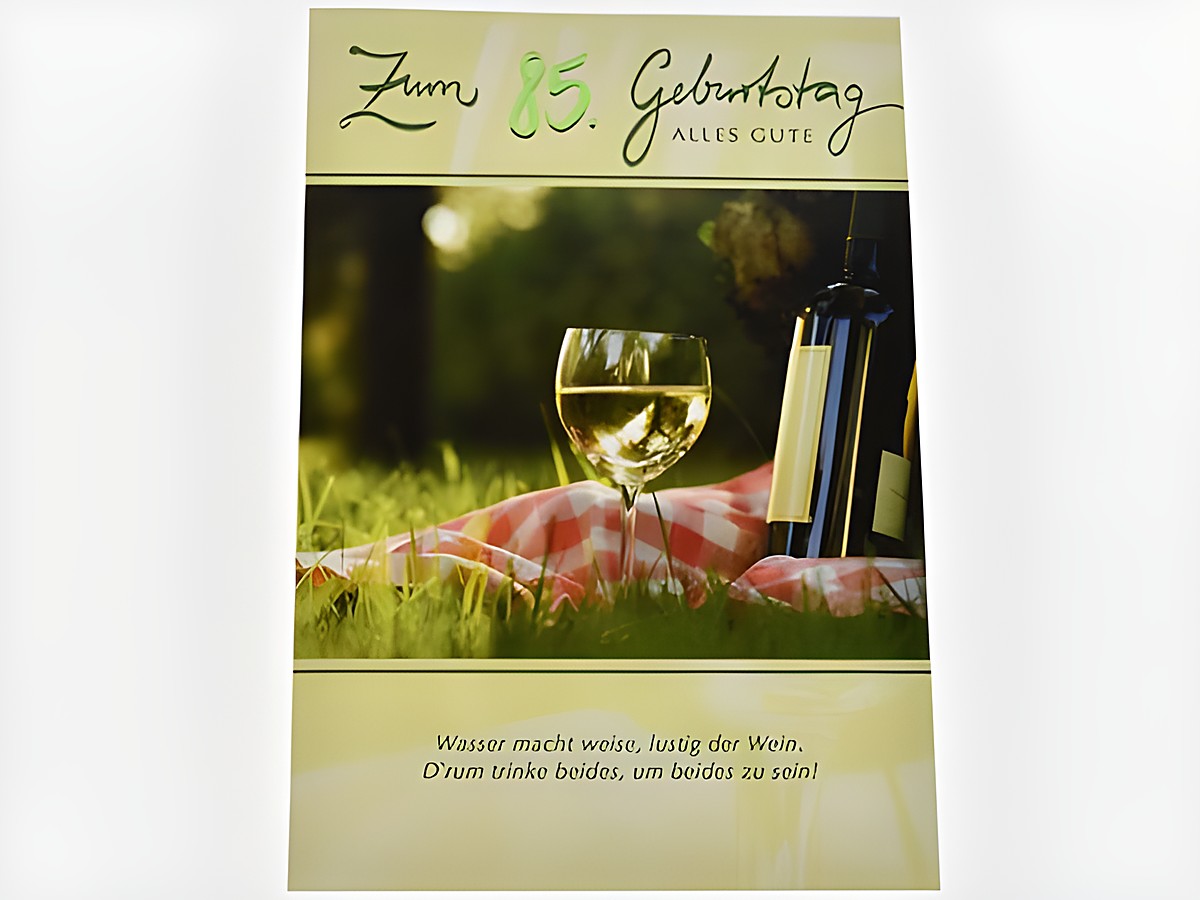 Eine Geburtstagskarte mit der Aufschrift zum 85. Geburtstag alles Gute, einer Flasche Wein und einem gefüllten Weinglas auf einer Picknickdecke im Grünen.