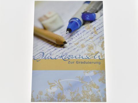 Auf dem Bild ist eine Glückwunschkarte zur Graduierung zu sehen, umgeben von Stiften und einem Schreibblock. Die Karte zeigt eine elegante Schrift und florale Motive.