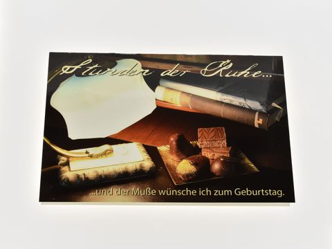 Geburtstagskarte mit dem Text Stunden der Ruhe und der Muße wünsche ich zum Geburtstag, dekoriert mit einer Lampe, Büchern und Pralinen, Hersteller Juwelier Harnisch.