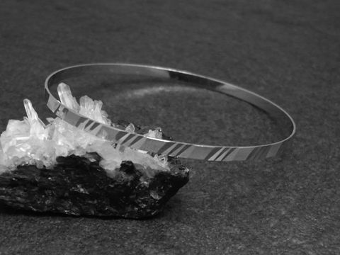 Zarter silberner Armreifen mit einem Durchmesser von 3,4 mm, rhodiniert, misst 21 cm und liegt auf einem schwarzen Untergrund neben transparenten Kristallen. Hersteller Juwelier Harnisch.