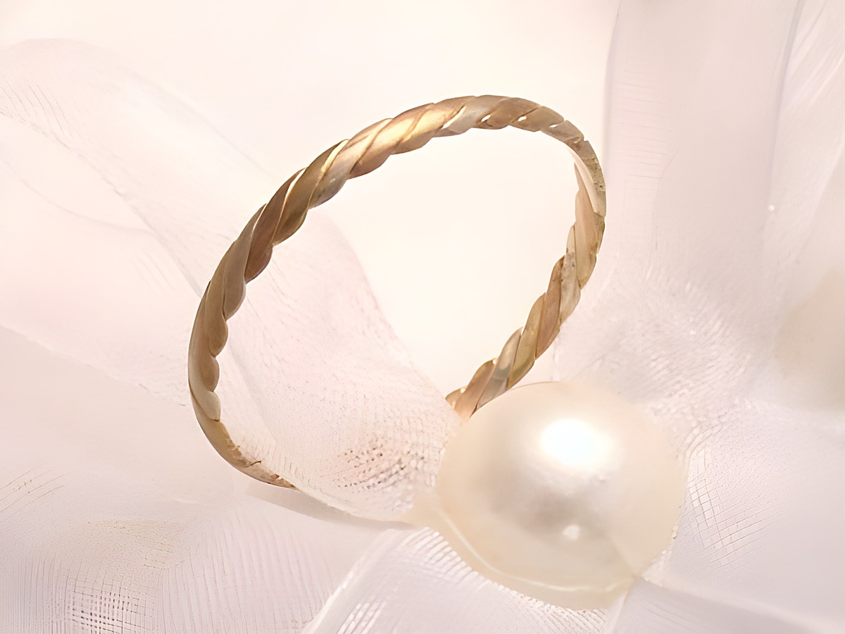 Trauring Ehering CEASELESS aus Gold 333 in Größe 58 und Durchmesser 18,4 mit gedrehter Oberfläche, vorgestellt auf einem weichen, hellen Untergrund zusammen mit einer Perle. Hersteller: Juwelier Harnisch.