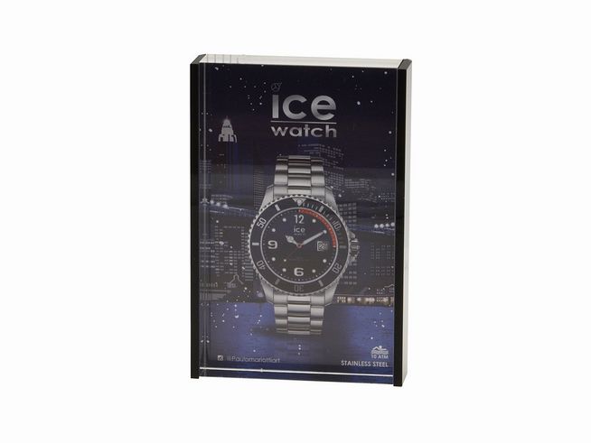 57350288-0 Ein schwarzer Bilderrahmen von Ice Watch zeigt eine stilvolle Armbanduhr mit Edelstahlarmband und schwarzem Ziffernblatt, platziert vor einer nächtlichen Stadtlandschaft.
