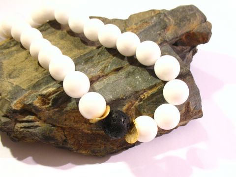 Eine elegante Kette aus weißen Opal-Kascholong-Perlen, ergänzt durch eine zentrale Lava-Perle und goldene Akzente, auf einem Stein platziert. Ideal für stilbewusste Schmuckliebhaber.