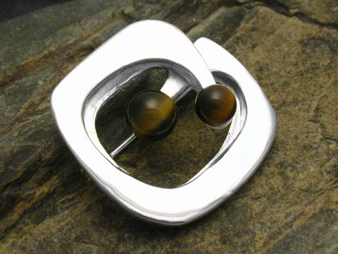Brosche Design mit Tigerauge echt Silber, Hersteller Juwelier Harnisch, elegante Form mit zwei runden Tigerauge-Steinen, auf einem rustikalen Untergrund präsentiert.