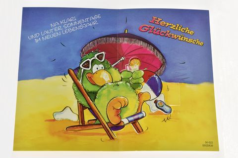 Bunte Geburtstagskarte mit einem fröhlichen Vogel, der auf einem Liegestuhl sitzt, Sonnenbrille trägt und Lottokugeln hält, farbenfroher Hintergrund mit Strandmotiv, Hersteller Juwelier Harnisch.