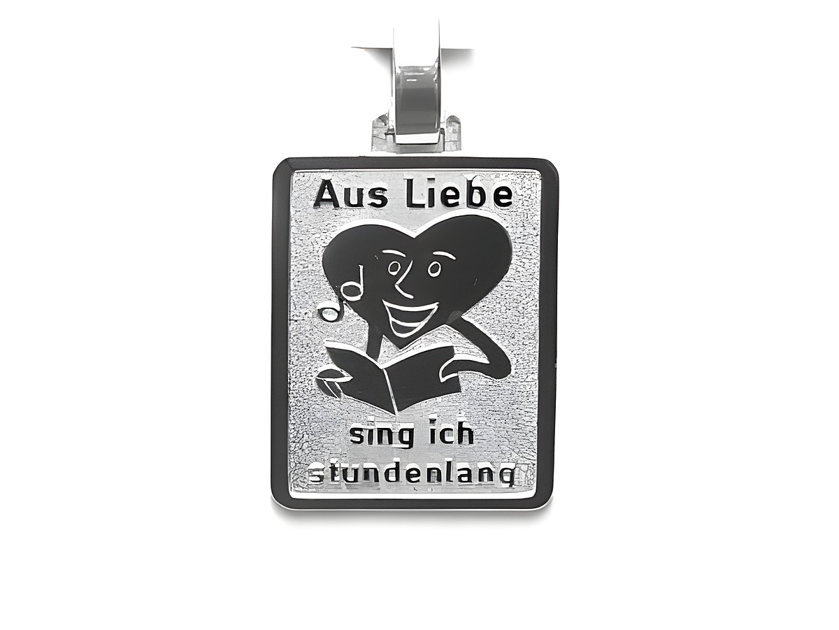 Silber Anhänger mit dem Motiv eines lachenden Gesichts, das eine herzförmige Figur umarmt. Darunter steht der Text Aus Liebe sing ich stundenlang. Hersteller Juwelier Harnisch.