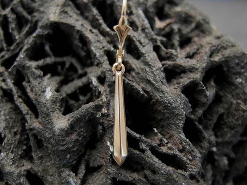 Ein eleganter unisex Ohrstecker aus goldfarbenem Material mit einer spitzen, verlaufenden Form, gefertigt von Juwelier Harnisch, präsentiert auf einer rustikalen Unterlage.
