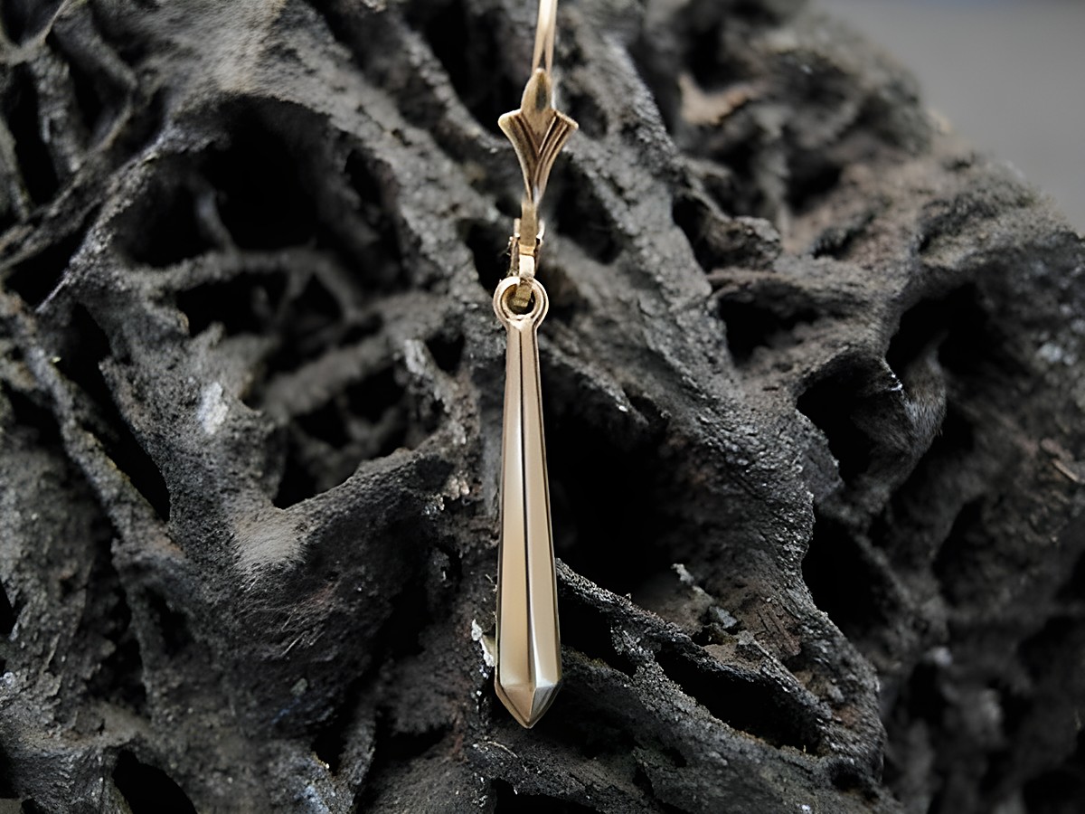 Ein eleganter unisex Ohrstecker aus goldfarbenem Material mit einer spitzen, verlaufenden Form, gefertigt von Juwelier Harnisch, präsentiert auf einer rustikalen Unterlage.