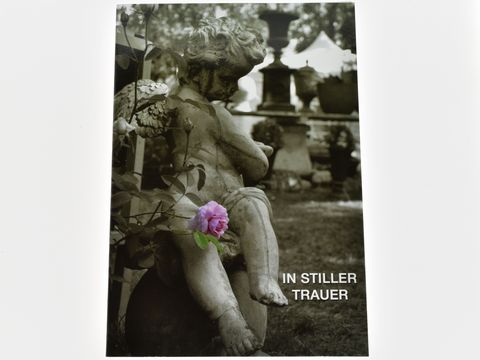Engel mit Rose, sitzend in nachdenklicher Pose. Im Hintergrund unscharfe Grabsteine. Text „IN STILLER TRAUER“ in weißer Schrift, insgesamt eine besinnliche, tröstende Atmosphäre.