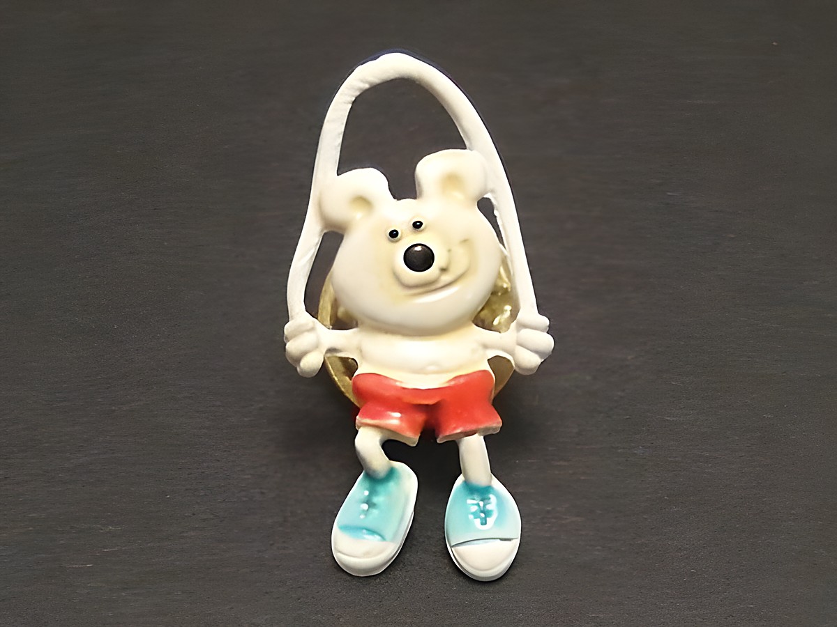 Bärchen mit Seil als Pin Anstecker von Juwelier Harnisch, der fröhliche Bär trägt rote Shorts und blaue Schuhe, ideal für eine verspielte Note auf Kleidung oder Taschen.