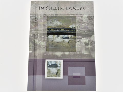 Eine Trauerkarte mit dem Text In stiller Trauer, abgebildet mit einer Szene von einer Brücke über ruhiges Wasser und Steinen, in sanften, dezenten Farben gestaltet, hergestellt von Juwelier Harnisch.