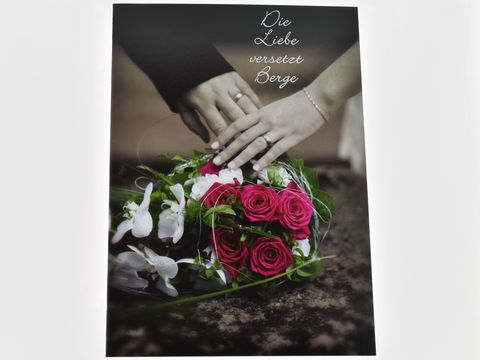 Auf der Hochzeitskarte sind zwei Hände zu sehen, die einen bunten Blumenstrauß halten. Der Strauß besteht aus roten Rosen und weißen Orchideen. Im Hintergrund steht der Text "Die Liebe versetzt Berge".