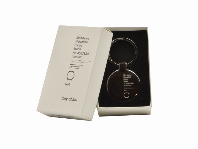 Ein Mondaine Helvetica Schlüsselanhänger aus Edelstahl liegt in einer weißen Box. Der Anhänger hat ein minimalistisches Design mit einem runden Kopf und einem Schlüsselring.
