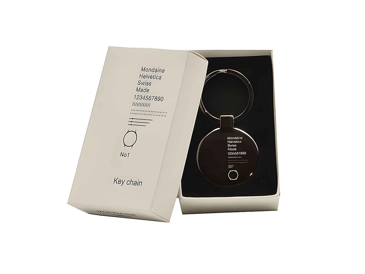 Ein Mondaine Helvetica Schlüsselanhänger aus Edelstahl liegt in einer weißen Box. Der Anhänger hat ein minimalistisches Design mit einem runden Kopf und einem Schlüsselring.