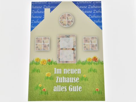 Ein stilisiertes Haus mit bunten Blumen auf einer Wiese, umgeben von grüner Rasenfläche. Darüber steht in blauer Schrift "Im neuen Zuhause alles Gute". Hersteller Juwelier Harnisch.