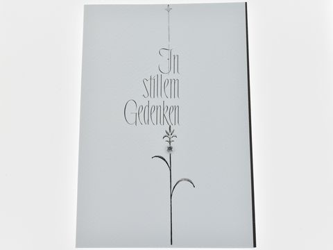 Weiße Trauerkarte mit strukturierter Oberfläche, beschriftet mit dem text In stillem Gedenken und einer stilisierten Blume. Hersteller Juwelier Harnisch, für Traueranlässe geeignet.