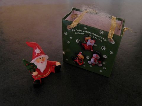 Weihnachtsmann mit Geschenktasche sitzt neben einem Tannenbaum, umgeben von einer grünen Geschenkebox mit Weihnachtsmotiven, Hersteller: Juwelier Harnisch, Weihnachten.