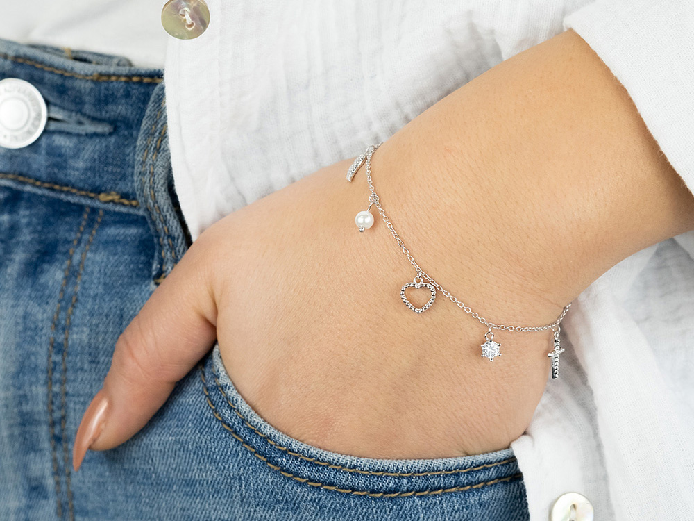 Silbernes Armband von Engelsrufer mit Zirkonia und Anhängern in Form eines Herzens, einer Perle und einem kleinen Kreuz, mit einer Länge von 17 plus 3 cm, an einem Handgelenk getragen.