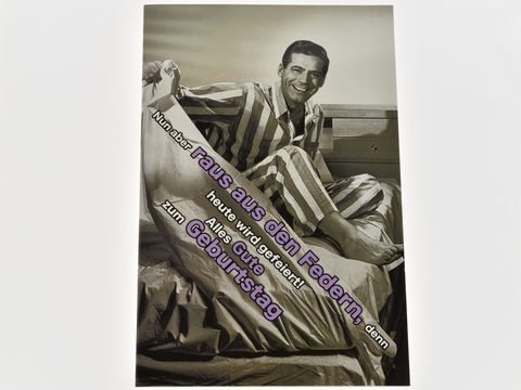 Eine Geburtstagskarte zeigt einen Mann im Bett, der in pyjamas gestreift ist. Er lächelt fröhlich und ist bereit, seinen Geburtstag zu feiern. Hersteller ist Juwelier Harnisch.